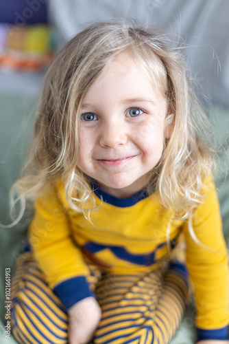 Cheerful toddler in cozy pajamas. Sitting happily and looking at the camera with bright blue eyes and long blond hair. Evoking feelings of love. Care. Selective focus