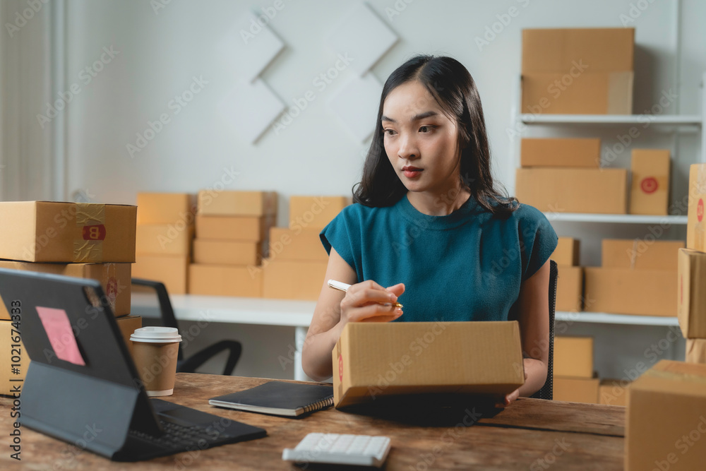 Young asian woman entrepreneur is writing a shipping address on a cardboard box while working in her home office Young asian woman entrepreneur is writing a shipping address on a cardboard box while working in her home office