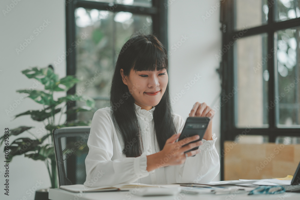 Young asian businesswoman happily smiles while browsing on her smartphone at her desk in a modern office, multitasking with paperwork and technology in a productive atmosphere Young asian businesswoman happily smiles while browsing on her smartphone at her desk in a modern office, multitasking with paperwork and technology in a productive atmosphere