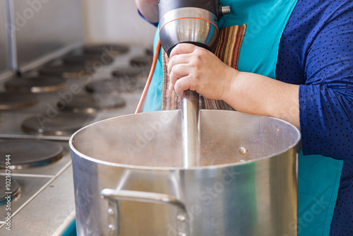 Cook is using an immersion blender while preparing a large amount of food in an industrial kitchen. Selective focus