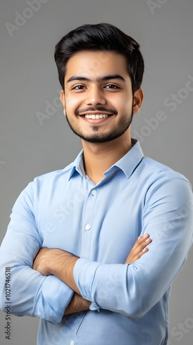 A portrait of an attractive young Indian man with wavy hair, smiling and standing confidently in front of a grey background, wearing a dark blue shirt