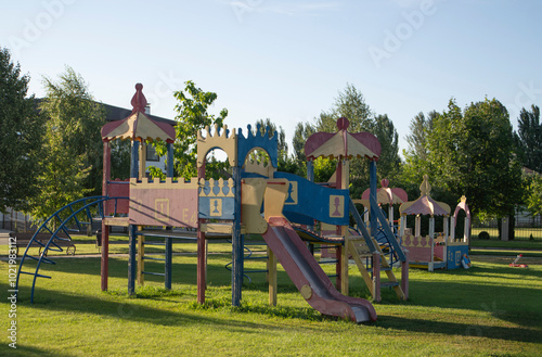 A vibrant playground features slides and climbing structures, surrounded by lush trees and grassy areas, where children enjoy playtime under the sun