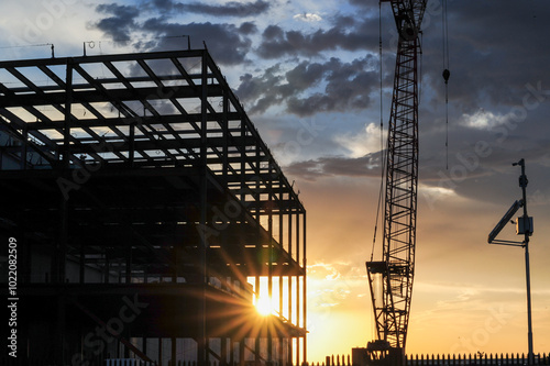 Obraz na plátně Sunset light outlines the steel beams and tower crane of a large commercial building under construction against a dark blue, cloudy sky
