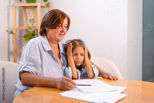 Shocked girl sitting with grandmother as they analyze financial bills on table at home. Little child is sitting with head in hands as the understand and manage their finances together 