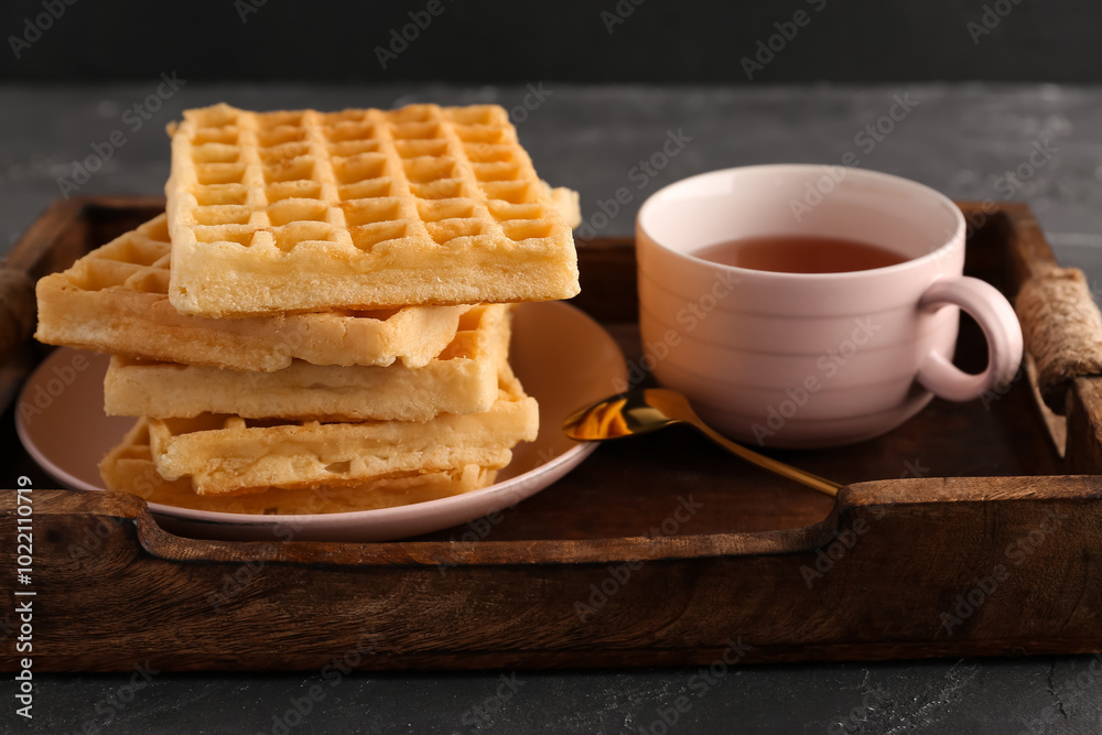 Wooden tray with tasty Belgian waffles and cup of tea on black background Wooden tray with tasty Belgian waffles and cup of tea on black background
