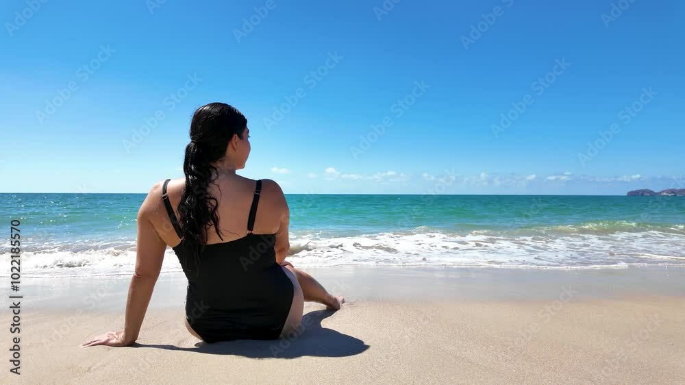 A young woman in a black swimsuit sits on the beach in San Carlos, Sonora, on a sunny day in front of the sea. She meditates as she enjoys this moment of introspection alone. A young woman in a black swimsuit sits on the beach in San Carlos, Sonora, on a sunny day in front of the sea. She meditates as she enjoys this moment of introspection alone.