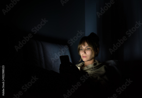 young woman staring at cell phone with face illuminated by screen late at night on sofa