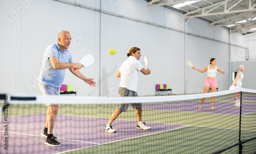 Portrait of concentrated experienced aged pickleball player preparing to strike and return ball to opponents field during doubles match in team with male partner on indoor court photo