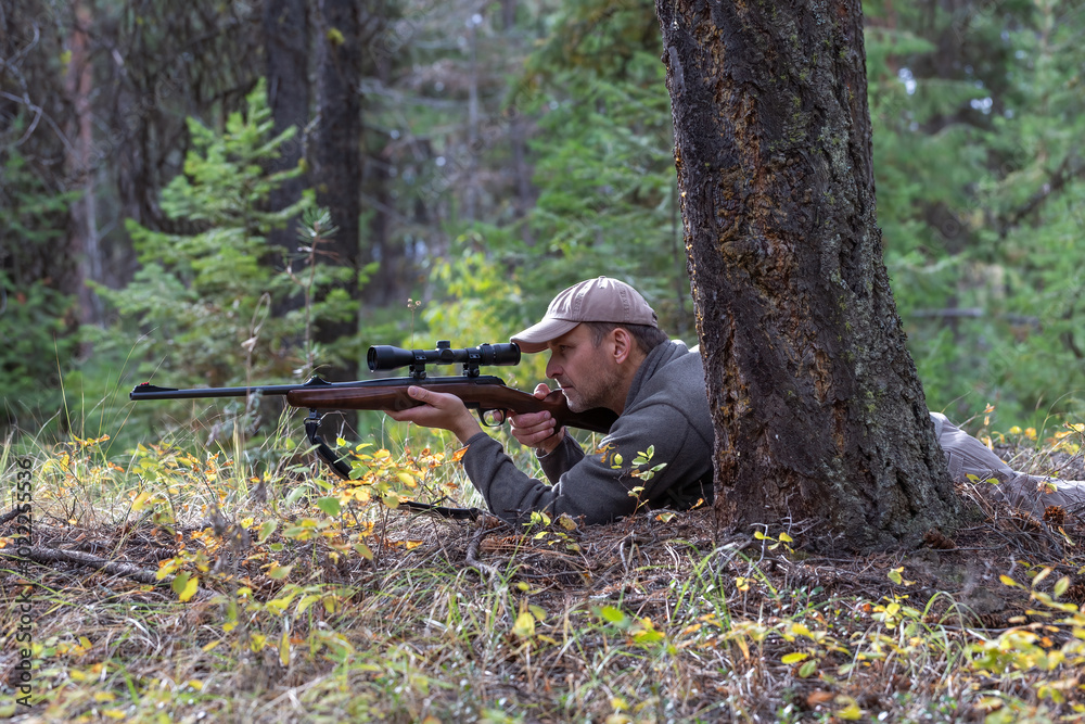 Naklejka premium Hunter lying on the ground, aiming his rifle at game while looking through the scope. Ready to shoot. Side view. Forest on background.