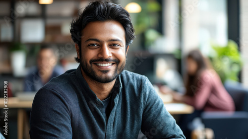 Casual Indian man in modern co working space smiling warmly, exuding confidence and approachability. vibrant atmosphere enhances feeling of collaboration and creativity