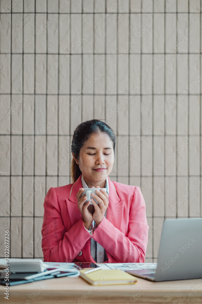 A professional woman in a pink blazer takes a moment to relax with a cup of coffee at her desk, surrounded by work materials and a laptop. A professional woman in a pink blazer takes a moment to relax with a cup of coffee at her desk, surrounded by work materials and a laptop.