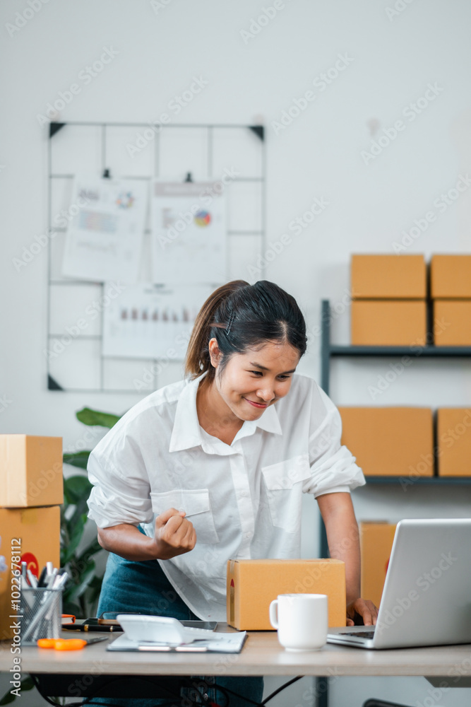A young businesswoman in a white shirt working on a laptop in a modern office with cardboard boxes and office supplies around her. A young businesswoman in a white shirt working on a laptop in a modern office with cardboard boxes and office supplies around her.