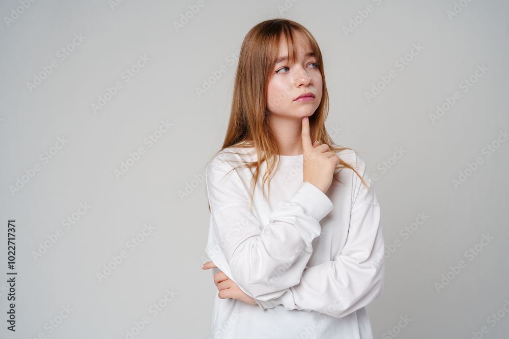 Young woman in a white long-sleeve shirt thoughtfully poses with a hand on her chin in a minimalistic setting Young woman in a white long-sleeve shirt thoughtfully poses with a hand on her chin in a minimalistic setting