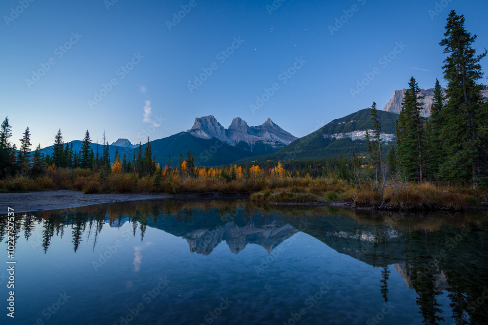 Fototapeta premium Beautiful scenery of the Canadian Rockies at dawn in autumn. Mountain, forest in yellow and green color reflected on water. Canmore, Alberta, Canada. The Three Sisters trio of peaks.