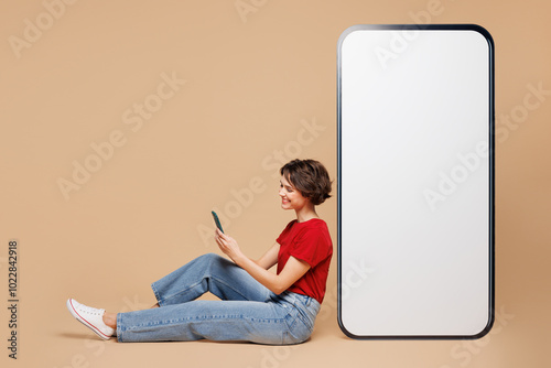 Full body side view young woman wear red t-shirt casual clothes sit near big huge blank screen mobile cell phone with area using smartphone isolated on plain light beige background. Lifestyle concept. photo