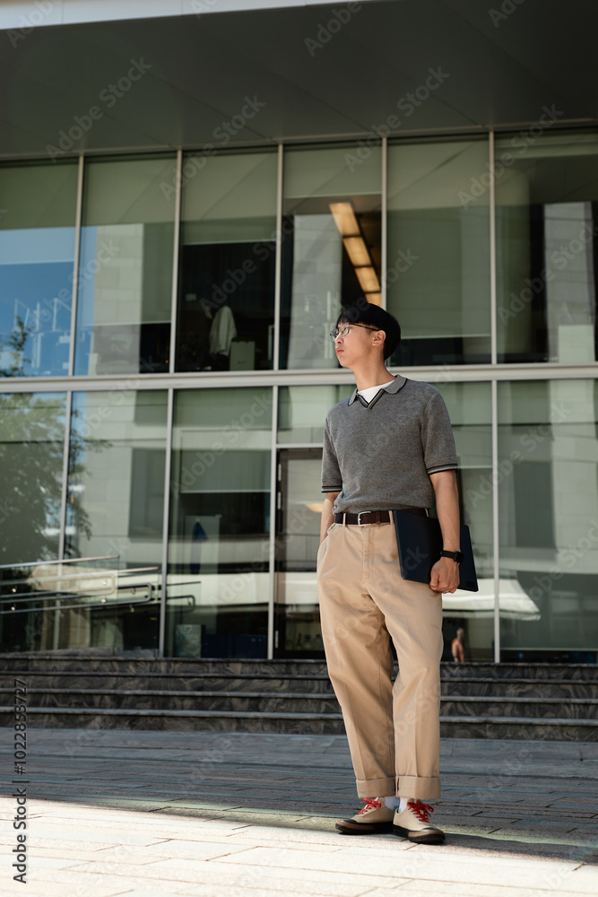 © Seventyfour - Vertical shot of fashionable Asian male IT programmer in office clothing holding laptop standing in sunlight next to business building while posing with hand in pocket outside in street, copy space