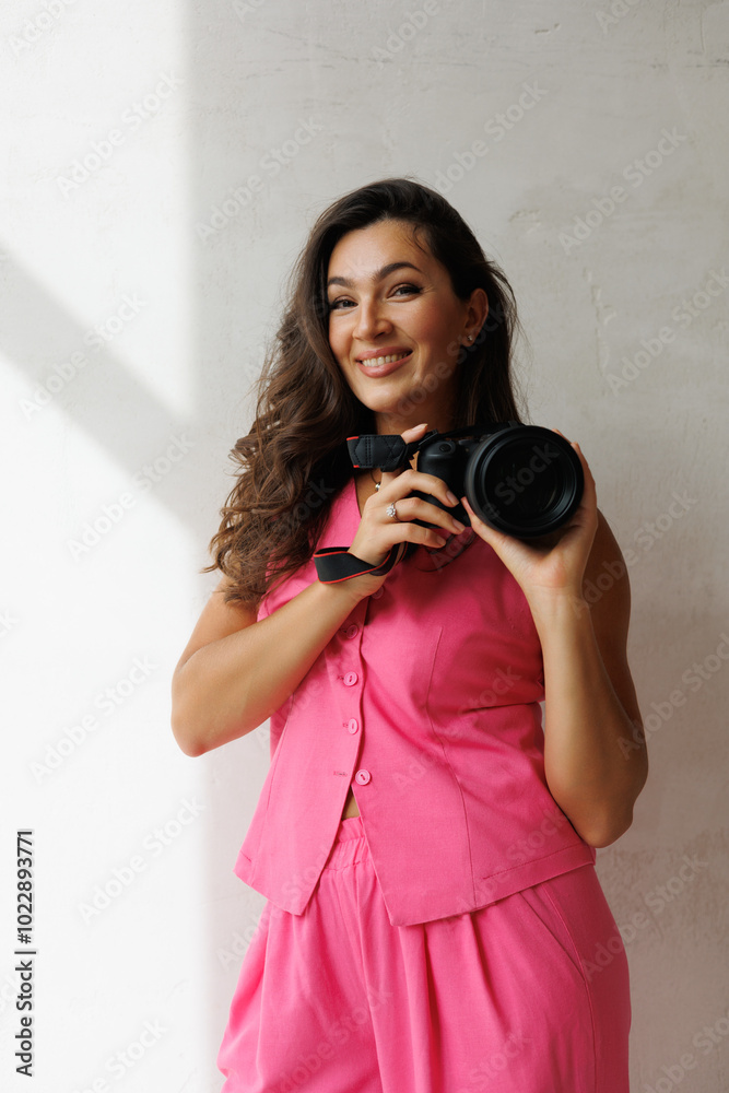 Obraz premium A beautiful tall brunette with spectacular styling stands at the window with a camera and smiles. A woman in a pink pantsuit in a vest and trousers holds a large camera and smiles. A photographer girl