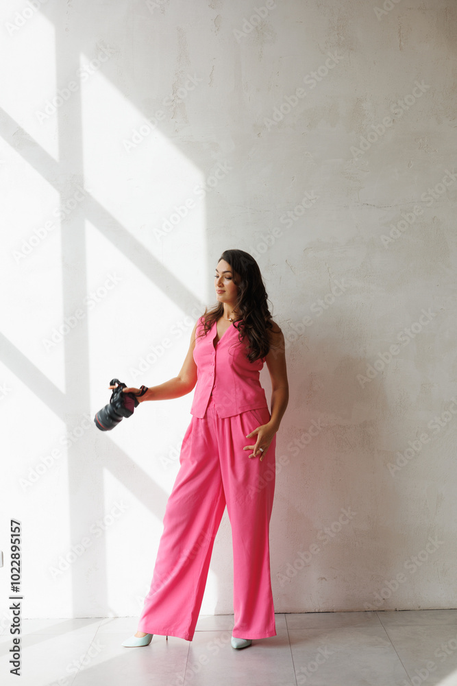 A beautiful tall brunette with spectacular styling stands at the window with a camera and smiles. A woman in a pink pantsuit in a vest and trousers holds a large camera and smiles. A photographer girl A beautiful tall brunette with spectacular styling stands at the window with a camera and smiles. A woman in a pink pantsuit in a vest and trousers holds a large camera and smiles. A photographer girl