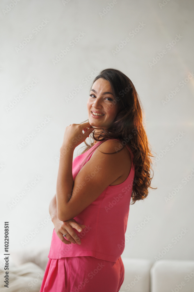 A beautiful tall brunette with spectacular styling stands at the window and smiles. A woman in a pink pantsuit in a vest and trousers on beige sofa. A beautiful tall brunette with spectacular styling stands at the window and smiles. A woman in a pink pantsuit in a vest and trousers on beige sofa.