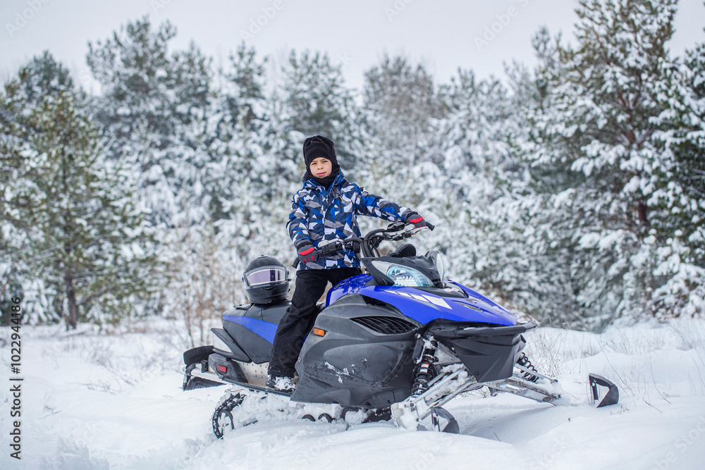 Boy driving snowmobile in a winter landscape. Little boy on the snowmobile up in the mountains smiling in a snowy weather, forest. Child rides a blue snowmobile on a snowy trail, pine trees Boy driving snowmobile in a winter landscape. Little boy on the snowmobile up in the mountains smiling in a snowy weather, forest. Child rides a blue snowmobile on a snowy trail, pine trees
