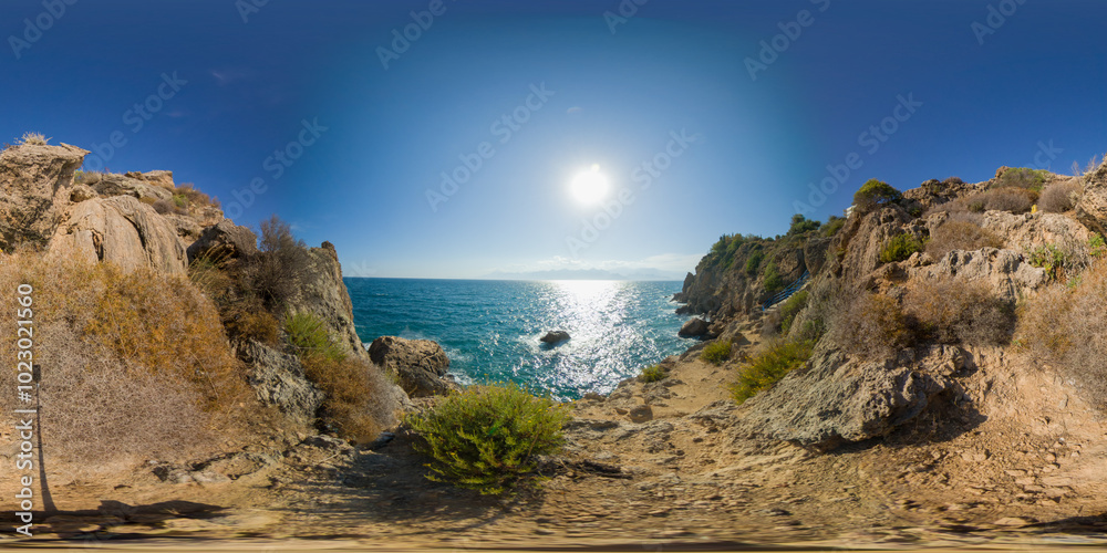 A rocky coastal path leading to the ocean with bright sunlight reflecting off the water. The 360-degree view captures rugged cliffs and natural vegetation along the shore. A rocky coastal path leading to the ocean with bright sunlight reflecting off the water. The 360-degree view captures rugged cliffs and natural vegetation along the shore.