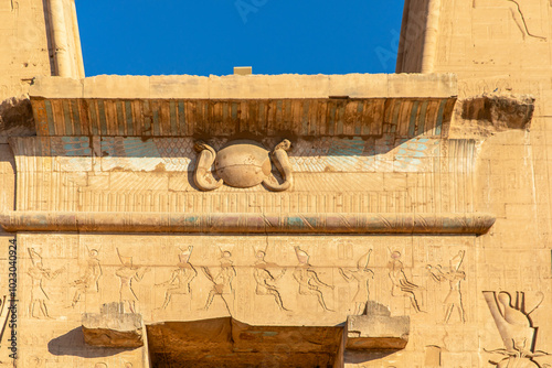 Soar through the heavens with the sun's wings over Edfu. The winged sun disk above the temple entrance symbolizes the divine power of Ra, illuminating Horus's sacred domain.