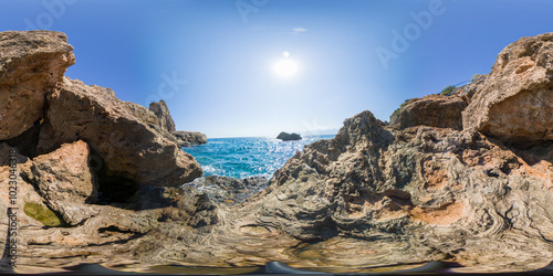 A rocky coastal scene with the bright sun shining over the ocean, captured in a 360-degree view. The rugged rocks and clear blue sky create a peaceful and scenic coastal environment.