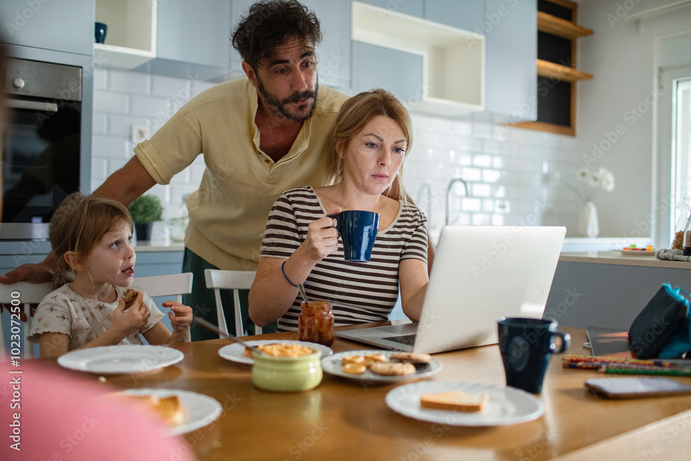 Mother working on laptop while family has breakfast in kitchen Mother working on laptop while family has breakfast in kitchen