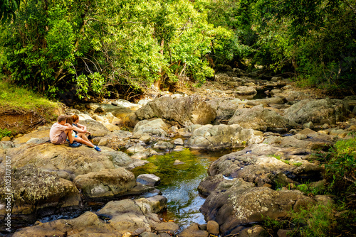 View of a family taking a break from hiking, sitting on a rock in the riverbed of a small stream that flows through the Vallée de Ferney in Mauritius