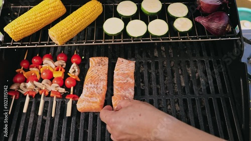 The chef carefully puts fresh salmon fillets on the grill, corn, zucchini and skewers of colorful vegetables..
