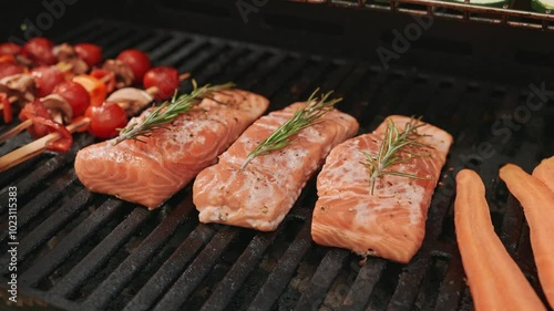 Close-up of salmon fillets topped with rosemary grilling on a barbecue, alongside skewers of cherry tomatoes and mushrooms.