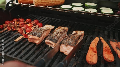 A chef's hands carefully place fillets of salmon, corn and vegetables on a grill for a vibrant outdoor cooking session.