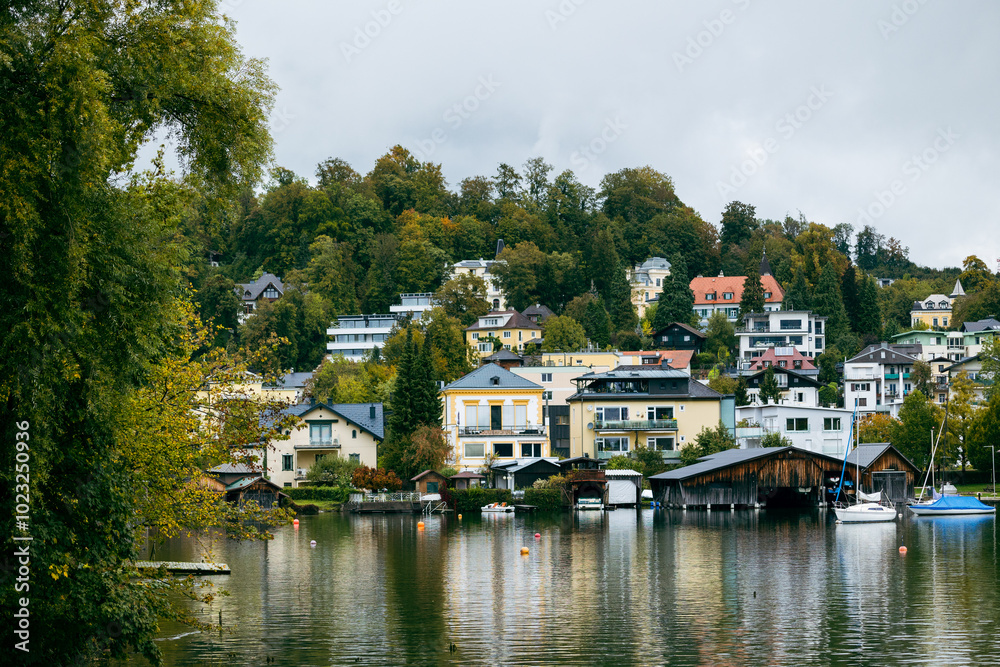 Fototapeta premium Colorful houses and buildings on the hillside by a lake, with reflections in the calm water. The scene captures a quaint lakeside village with a peaceful and picturesque atmosphere