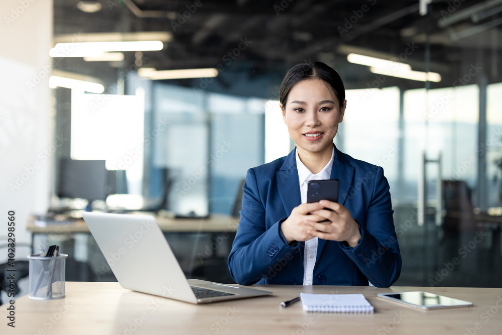 Asian business woman in office using smartphone, sitting at desk with laptop, notebook. Professional setting, confident expression. Concepts include technology, communication, business, work. Asian business woman in office using smartphone, sitting at desk with laptop, notebook. Professional setting, confident expression. Concepts include technology, communication, business, work.