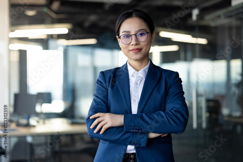 Asian business woman confidently standing in office, arms crossed, wearing glasses and suit. Emphasizes professionalism, leadership, corporate success, female empowerment in modern workplace.