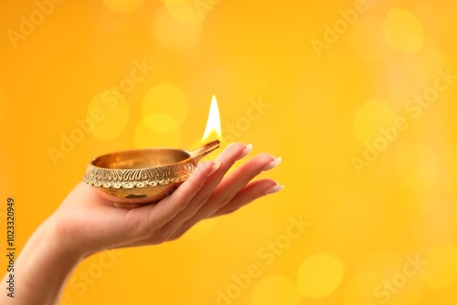 Diwali celebration. Woman holding lit diya lamp on yellow background with blurred lights, closeup. Space for text photo