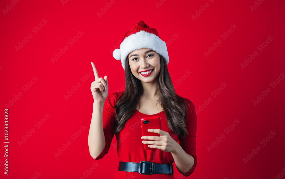 Attractive young woman in red dress and Santa Claus hat holding red smartphone on red festive background and pointing finger. Hand gesture attracting attention. Attractive young woman in red dress and Santa Claus hat holding red smartphone on red festive background and pointing finger. Hand gesture attracting attention.