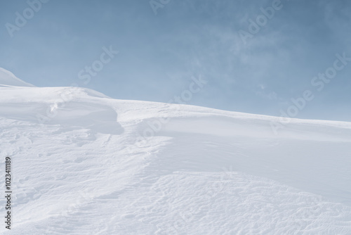 A vast, undisturbed expanse of snow stretches beneath a clear, blue sky. Gentle ridges and subtle shadows create a sense of peace and tranquility.