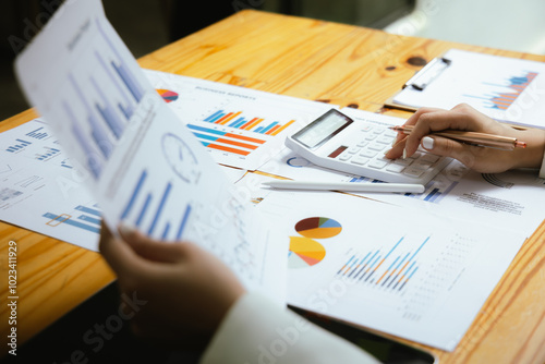 A businesswoman sits at her desk, calculating financial data on her laptop. Surrounded by paperwork and charts, she analyzes expenses and income, reflecting her role in corporate finance and accountin photo