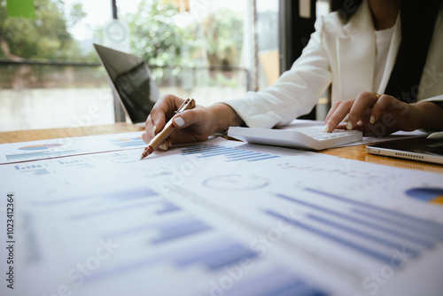 A businesswoman sits at her desk, calculating financial data on her laptop. Surrounded by paperwork and charts, she analyzes expenses and income, reflecting her role in corporate finance and accountin photo