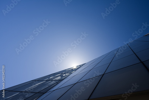 A modern glass building angles upwards towards a clear, blue sky with the sun near its peak.