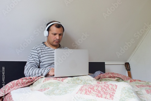 latino man with headphones, is sitting on a bed with a laptop computer.