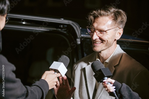 Medium shot of smiling male celebrity in camera light speaking with TV media enjoying public eye attention while journalists taking superstars exclusive comment in night event, copy space photo