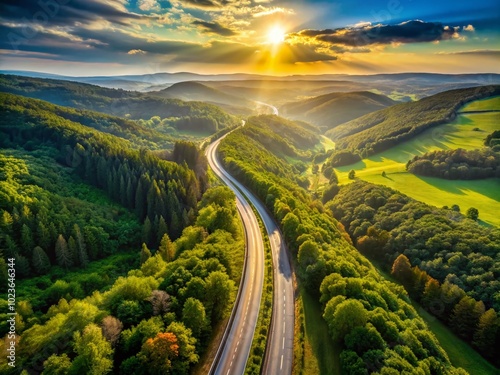 Aerial perspective of a winding highway surrounded by lush green forests and rolling hills, stretching into the distance towards a sun-kissed horizon. photo