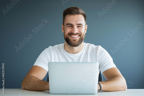 Smiling man in casual attire sitting at a desk with a laptop during a bright workday