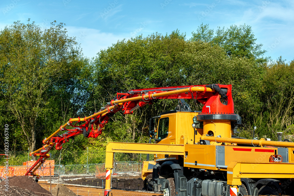 Bright yellow mobile concrete pump pouring concrete on a sunny day surrounded by lush green trees