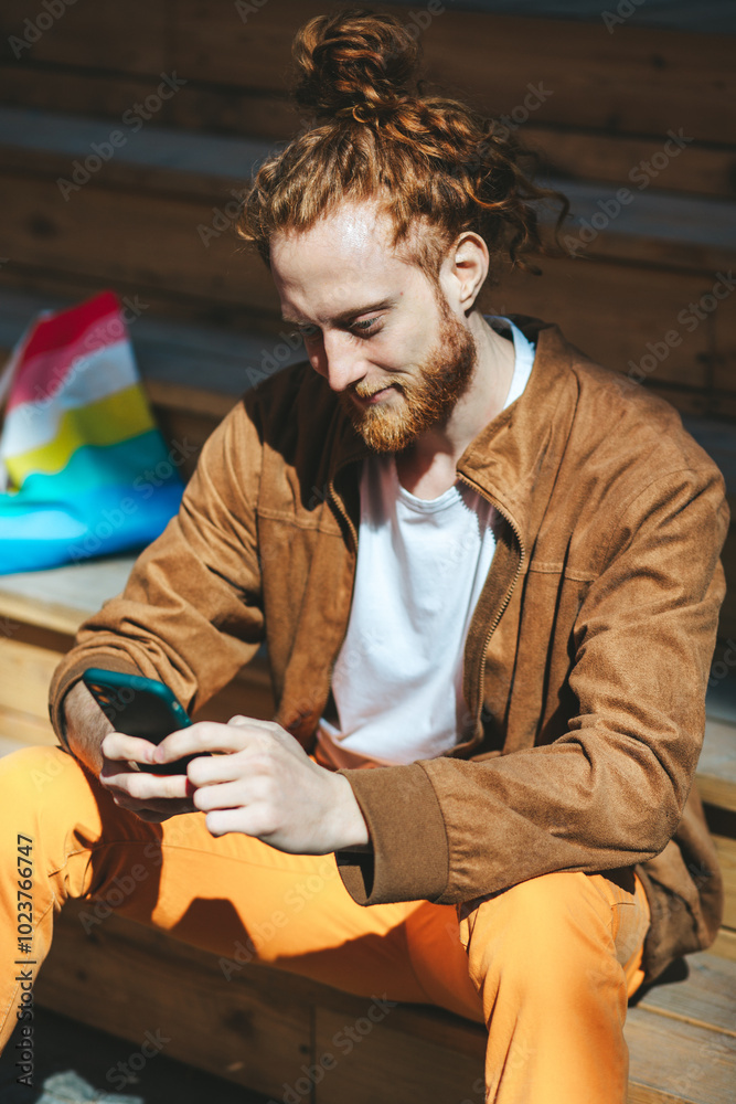 Young man with fiery red hair and a well groomed beard sits on weathered steps, using his smartphone on a sunny day. He looks content, embodying the connection between tech and urban life