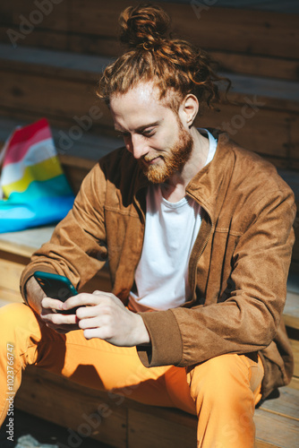 Obraz na płótnie Young man with fiery red hair and a well groomed beard sits on weathered steps, using his smartphone on a sunny day. He looks content, embodying the connection between tech and urban life w salonie