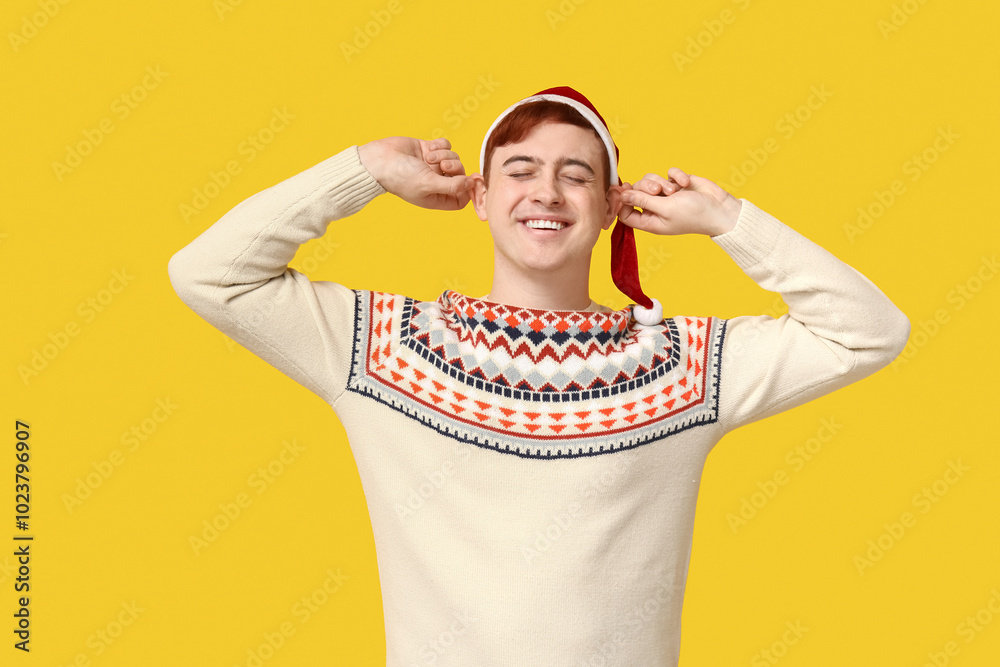 Handsome young man in warm sweater and Santa hat on yellow background Handsome young man in warm sweater and Santa hat on yellow background