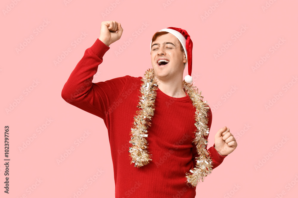 Handsome young man in warm sweater, Santa hat and tinsel on pink background Handsome young man in warm sweater, Santa hat and tinsel on pink background